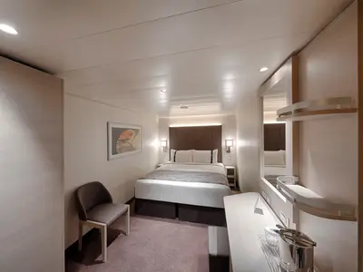 Eine moderne Innenkabine an Bord der MSC World Asia. Zu sehen sind ein gemachtes Doppelbett mit Kopfteil, Nachttische, eine kleine Sitzgelegenheit und ein Spiegelbereich mit Ablagen.