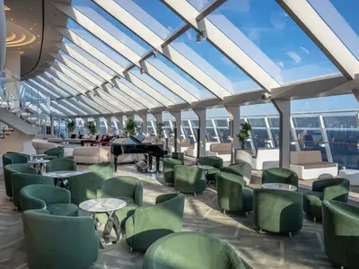 Helle, elegante Lounge der MSC World Asia mit grossen Fenstern und Glasdach. Grüne Sessel, weisse Sofas, ein Flügel und Meerblick prägen den modernen Raum.