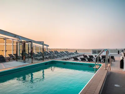 Der Zen-Pool an Bord der MSC World Asia am späten Nachmittag. Türkisfarbenes Poolwasser reflektiert den orangefarbenen Himmel über dem Meer. Zahlreiche Liegestühle stehen bereit.