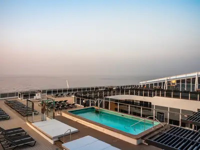 Blick über das ruhige Zen-Pool-Deck an Bord der MSC World Asia bei Sonnenaufgang. Mehrere leere Sonnenliegen sind um den blau leuchtenden Pool angeordnet. Im Hintergrund erstreckt sich das weite, ruhige Meer bis zum Horizont unter einem pastellfarbenen Himmel.