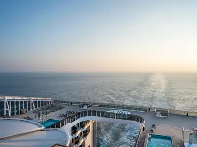 Blick vom Heck der MSC World Asia auf das offene Meer während des Sonnenaufgangs. Das weitläufige Sonnendeck mit Pools und zahlreichen Sonnenliegen ist im Vordergrund zu sehen.