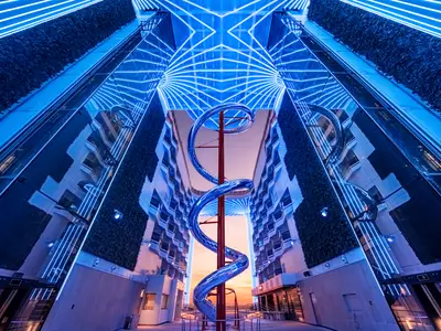 Die futuristische World Promenade an Bord der MSC World Asia bei Sonnenuntergang. Eine hohe blaue Spiralrutsche ist zentral. Blaue Lichtinstallationen an Dach und Wänden spiegeln sich im Glas. Grüne Gärten und Balkone säumen den Raum.
