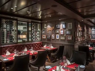 Innenansicht des eleganten Steakhouse "Butcher's Cut" an Bord der MSC World Asia, mit rot gepolsterten Sitzbänken, dunklen Holztischen und einer dekorativen Wand mit gerahmten Bildern und einem Rindfleisch-Schnittmuster. Eine beleuchtete Weinauslage ist links zu sehen.
