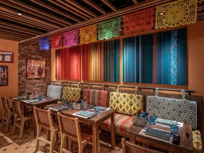 Das Hola! Tacos & Cantina Restaurant der MSC World Asia zeigt Holztische und Sitzbänke mit bunten Kissen. Die Wände sind mit Streifenkunst und mexikanischen Bannern verziert. Eine große Kunstcollage hängt an einer Backsteinwand, was eine lebhafte und einladende Atmosphäre schafft.
