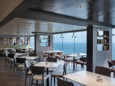 Ein helles und modernes Restaurant an Bord der MSC World Asia mit raumhohen Fenstern, die einen weiten Blick auf das Meer bieten. Weiße Tische und dunkle Stühle füllen den Raum, ergänzt durch eine lange grüne Sitzbank und dekorative Regale an den Wänden.