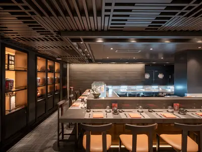 Innenansicht des Kaito Sushi Bar Spezialitätenrestaurants an Bord der MSC World Asia. Es zeigt eine lange Theke mit vorbereiteten Plätzen, einer offenen Küche im Hintergrund und beleuchteten Regalen mit Dekorationen.