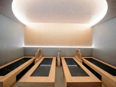 Moderner Ganbanyoku-Bereich im MSC Aurea Spa an Bord der MSC World Asia. Helle Holzliegen mit dunklen Marmorflächen sind in einem minimalistischen Raum mit indirekter Beleuchtung und einer Mischung aus hellem Holz und grauen Fliesen zu sehen. Die Szene ist beruhigend und ohne Personen.