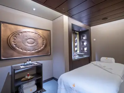 Ein eleganter Spa-Behandlungsraum an Bord der MSC World Asia. Eine weisse Massageliege und ein kunstvolles Wandrelief dominieren den Raum. Hinten ein Waschbeckenbereich.