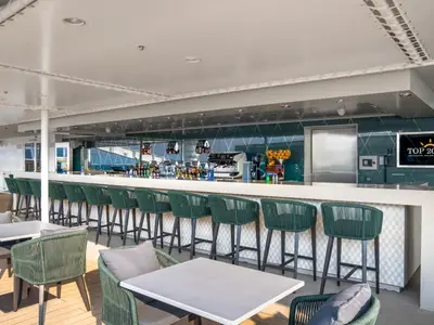 Moderne Bar mit weisser Theke und grünen Barhockern an Bord der MSC World Asia. Im Hintergrund spiegeln sich Himmel und Meer an der Wandverkleidung wider. Der Bereich ist überdacht.