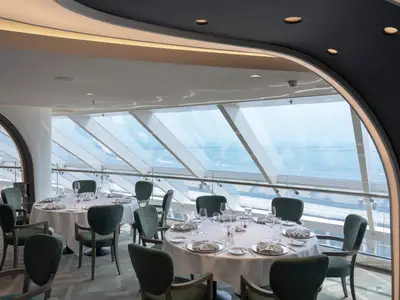 Ein elegantes Spezialitätenrestaurant an Bord der MSC World Asia mit stilvoll gedeckten Tischen und dunkelgrünen Stühlen. Große Glasfenster bieten einen weiten Blick auf das offene Meer. Der Raum ist hell und modern gestaltet.