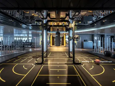 Das moderne Fitnessstudio an Bord der MSC World Asia zeigt einen grossen Raum mit verschiedenen Trainingszonen. Im Vordergrund ein Bereich mit gelben Bodenmarkierungen und einer Hantelstange. Dahinter eine Multifunktionsstation mit Boxsack, umgeben von gelben Säulen. Im Hintergrund sind weitere Fitnessgeräte zu sehen.