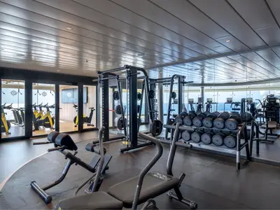 Moderner Fitnessraum an Bord der MSC World Asia mit Hantelregalen, Kraftgeräten und stationären Fahrrädern. Große Fenster bieten einen weiten Meerblick. Der Boden ist mit dunklen Sportmatten ausgelegt.