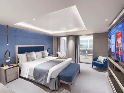 Eine helle, moderne Suite an Bord der MSC World Asia. Sie zeigt ein Kingsize-Bett, eine blaue Akzentwand, Sitzbank und Sessel. Ein großer Fernseher ist rechts sichtbar, Nachttisch links. Eine Glastür führt zum privaten Balkon mit Meerblick.