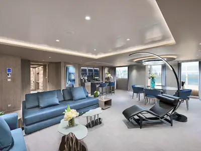 Blick in die helle und moderne MSC Yacht Club Owner's Suite der MSC World Asia mit blauen Sofas, Bar, Esstisch und Meerblick.
