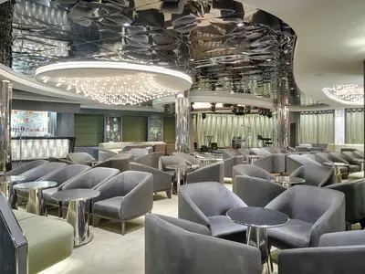 Elegante Dolce Vita Bar an Bord der MSC World Atlantic mit vielen grauen Lounge-Sesseln, Tischen, einer Bar und Bühne mit Instrumenten. Auffällige Deckenbeleuchtung.