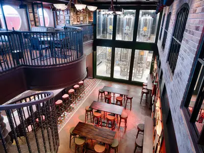 Vogelperspektive der Masters Of The Sea Bar an Bord der MSC World Atlantic. Der mehrstufige Barbereich mit Tischen, Hockern und einer Treppe ist zu sehen. Glänzende Biertanks der Bordbrauerei sind hinter großen Fenstern sichtbar.