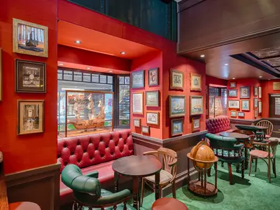 Blick in die detailreich dekorierte Masters Of The Sea Bar an Bord der MSC World Atlantic. Rote Waende und Ledersofas, gruener Teppich und maritime Bilder sowie ein Schiffsmodell schmuecken den Raum.
