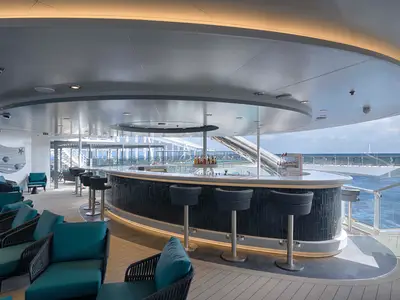 Die moderne Zen Bar an Bord der MSC World Atlantic. Überdachter Bereich mit Bar, Hockern, Loungemöbeln und Meerblick. Wasserrutsche im Hintergrund.