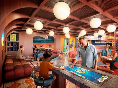 Eine moderne, farbenfrohe Lounge an Bord der MSC World Atlantic. Mehrere Personen sind zu sehen, darunter ein Vater und Sohn, die an einem interaktiven Spieletisch spielen. Im Hintergrund sitzen weitere Gäste an Tischen oder auf Sofas und unterhalten sich. Die Einrichtung ist hell mit Holzelementen und grossen, runden Deckenlampen.