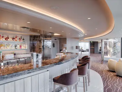 Moderne Bar-Lounge an Bord der MSC World Atlantic mit langer Bartheke, Hockern und Regalen voller Spirituosen. Indirekte Beleuchtung und gemusterter Teppich. Ohne Personen.