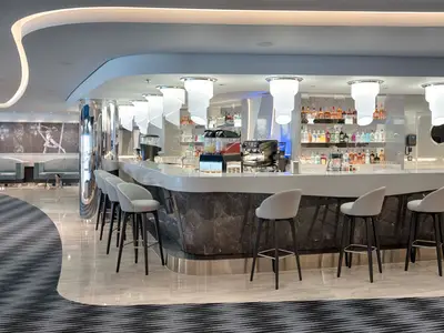 Die Bar-Lounge an Bord der MSC World Atlantic zeigt einen geschwungenen Bartresen mit hellen Hockern und Spirituosenregalen. Hinten: Sofas und Wandkunst.