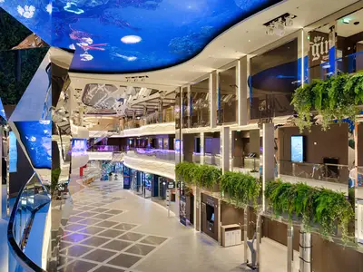 Die World Galleria an Bord der MSC World Atlantic. Eine moderne, mehrstöckige Promenade mit Geschäften und Hängepflanzen. Ein großer LED-Deckenbildschirm zeigt eine Unterwasserwelt.
