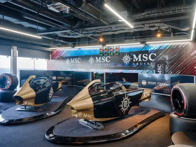 Zwei gold-schwarze Rennsimulatoren in der Formel-Racer-Zone an Bord der MSC World Atlantic. Bildschirme zeigen Markenlogos, Startampeln und Rennreifen dekorieren. Meerblick durch Fenster. Ohne Personen.