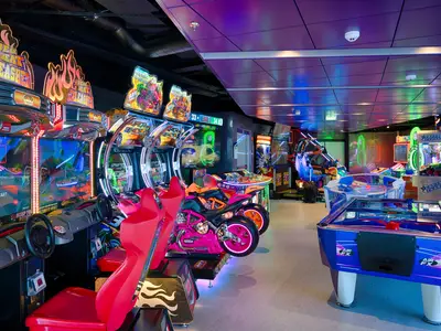 Eine farbenfrohe und lebhafte Spielhalle an Bord der MSC World Atlantic mit zahlreichen Arcade-Spielen, darunter Rennsimulator-Automaten und Airhockey-Tische, beleuchtet von Neonlichtern. Keine Personen sichtbar.