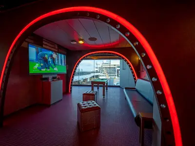 Junior Club an Bord der MSC World Atlantic. Rote LED-Bögen, TV mit Fußball, Konsolen, Billardtisch. Fenster zeigen Meerblick und anderes Schiff. Dunkle Sitze.