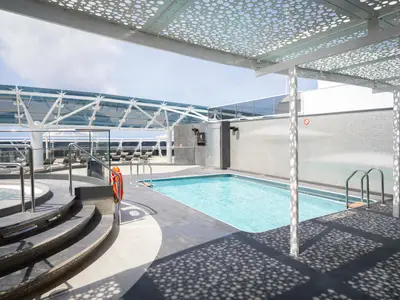 Poolbereich des MSC Yacht Clubs an Bord der MSC World Atlantic. Helles Schwimmbecken, Whirlpool, moderne Pergola mit Lochmuster. Erzeugt Schatten auf dem Deck. Liegestühle im Hintergrund.