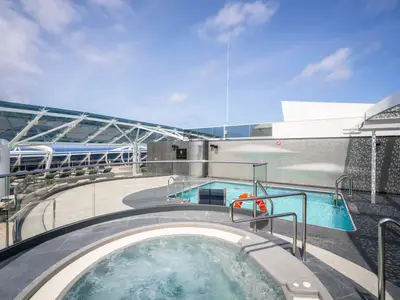 Der moderne Pool- und Wellnessbereich an Deck der MSC World Atlantic. Im Vordergrund ein sprudelnder Jacuzzi, dahinter ein länglicher Pool. Sonnenliegen, eine transparente Glasüberdachung und weite Meerblicke unter blauem Himmel sind zu sehen.