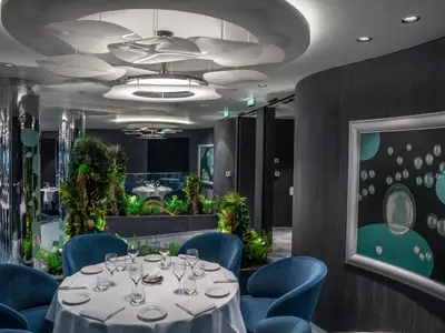 Modernes Bubbles Spezialitätenrestaurant der MSC World Atlantic. Blaue Stühle, weiße Tische, Pflanzen, glänzende Säulen. Kunstinstallation mit Blasenmotiven. Ohne Personen.
