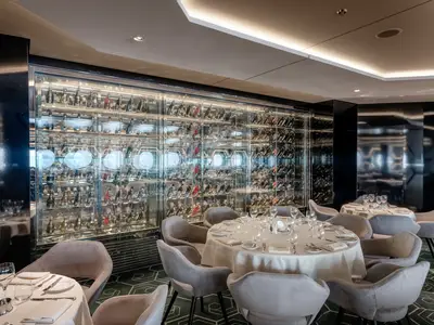 Innenansicht des Restaurants Esagono an Bord der MSC World Atlantic. Elegante Tische und Stühle stehen vor einer grossen, beleuchteten Glaswand mit vielen Weinflaschen. Modernes Interieur in Grautönen.