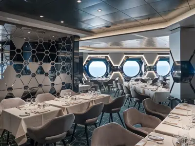 Restaurant Esagono an Bord der MSC World Atlantic mit gedeckten Tischen und bequemen Stühlen. Große Bullaugen zeigen Meerblick. Moderner Innenraum mit auffälligen sechseckigen Wandelementen.