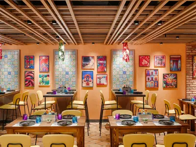 Ein farbenfrohes Spezialitätenrestaurant, Hola! Tacos & Cantina, an Bord der MSC World Atlantic. Die Wände sind orange mit gemusterten Fliesen und mexikanisch inspirierten Bildern. Holzdeckenbalken sind sichtbar. Im Vordergrund stehen Holztische mit schwarzen Tellern und bunten Gläsern, umgeben von gelben Stühlen.