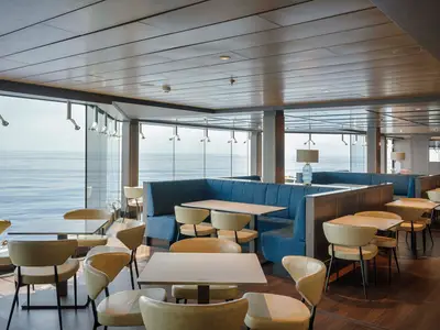 Ein modernes Restaurant an Bord der MSC World Atlantic. Blaue Polsterbänke und helle Stühle an Tischen bieten Platz mit Panoramablick auf das offene Meer.