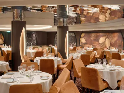 Ein elegantes Restaurant an Bord der MSC World Atlantic mit zahlreichen runden Tischen, eingedeckt mit weissen Tischdecken, Geschirr und Gläsern. Samtbezogene Stühle in warmem Orangeton stehen um die Tische. Die Wände sind mit grossflächigen Kunstwerken von goldenen Blättern verziert. Eine wellige, spiegelnde Decke reflektiert das Licht.