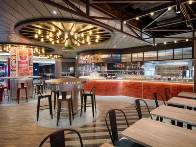 Ein lebhaftes, modernes Fast-Casual-Restaurant an Bord der MSC World Atlantic. Im Vordergrund sind Tische und Barhocker, dahinter eine Pizzastation mit Auslage und roten Fliesen zu sehen. An einer Säule sind Burger- und Pommes-Motive.