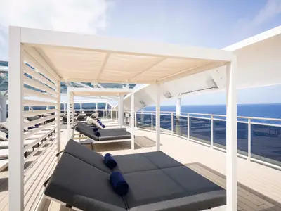 Sonnendeck an Bord der MSC World Atlantic mit Liegestühlen und Pergolen. Blaue Handtücher liegen bereit. Weitblick auf das blaue Meer und den klaren Himmel durch eine Glasbrüstung.
