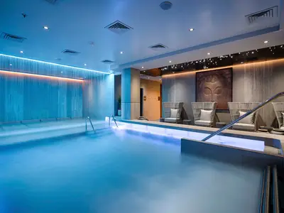 Der hellblau beleuchtete Innenpool des MSC Aurea Spa an Bord der MSC World Atlantic. Eine Wasserwand und Ruhezone mit Korbsesseln und Buddha-Skulptur.