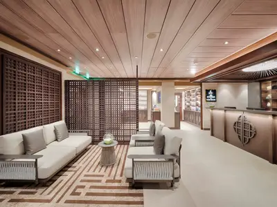Der elegante Empfangsbereich des MSC Aurea Spa an Bord der MSC World Atlantic mit dunklen Holzgittern, modernen weissen Sofas und einem Rezeptionstresen. Links sind Produktauslagen zu sehen.
