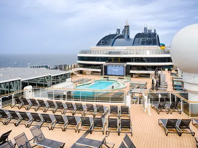 Pooldeck der MSC World Atlantic mit Sonnenliegen, blauem Pool, Bühne (MSC-Logo) und Glasbauten. Rechts großer Radom. Dahinter offenes Meer unter bewölktem Himmel.