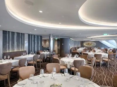Innenansicht des eleganten Spezialitätenrestaurants an Bord der MSC World Atlantic. Zahlreiche Tische mit weißen Tischtüchern, Gläsern und Besteck. Beige Stühle, dunkle Sitzbänke und modernes Wanddesign prägen den gehobenen Raum.