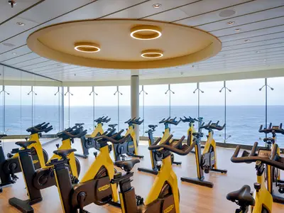 Helles und modernes Fitnessstudio an Bord der MSC World Atlantic. Viele gelb-schwarze Spinning-Räder stehen vor großen Fenstern mit Panoramablick auf das Meer. Runde Deckenleuchten.