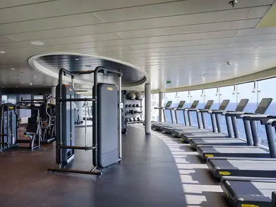 Ein breiter Blick in den geräumigen Fitnessbereich an Bord der MSC World Atlantic. Links stehen Krafttrainingsgeräte, rechts reihen sich Laufbänder entlang einer großen Fensterfront mit Blick auf das offene Meer unter blauem Himmel. Der Raum ist hell und modern eingerichtet.