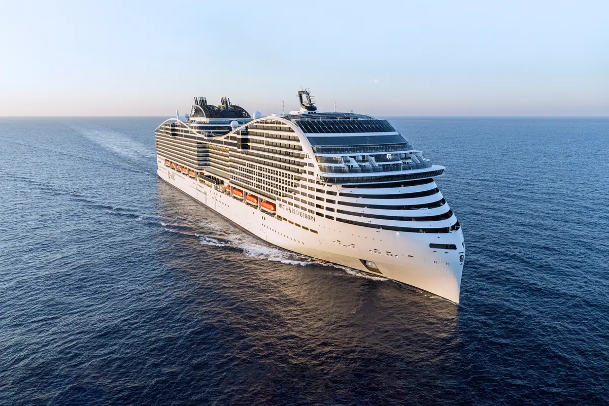 Luftaufnahme der MSC World Europa, einem riesigen weissen Kreuzfahrtschiff, das auf dem offenen Meer fährt. Man sieht die Steuerbordseite, Bugwelle im tiefblauen Wasser, klarer Himmel.