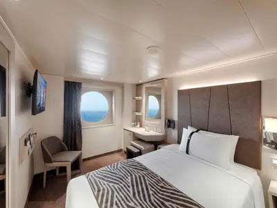 Eine modern eingerichtete Aussenkabine an Bord der MSC World Europa. Ein Bullauge bietet Meerblick. Zu sehen sind ein Doppelbett, ein Schminktisch mit Spiegel und ein Sessel. Das Zimmer ist hell beleuchtet.