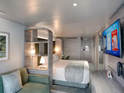 Blick in eine moderne Balkonkabine an Bord der MSC World Europa. Ein Doppelbett, eine grüne Couch und ein Flachbildfernseher sind zu sehen. Helle Holz- und Grautöne dominieren das Interieur.
