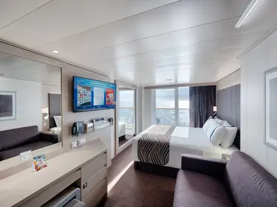Geräumige und moderne Balkonkabine an Bord der MSC World Europa mit Doppelbett, Sofa, TV und Meerblick vom privaten Balkon aus. Tageslicht fällt in den Raum.