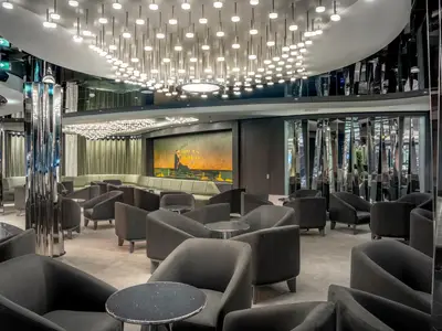 Blick in die elegante Dolce Vita Bar an Bord der MSC World Europa. Dunkle Sessel und glänzende Tische stehen vor einer langen Sitzbank. Eine kunstvolle Deckenbeleuchtung und ein großes Wandgemälde prägen den Raum.